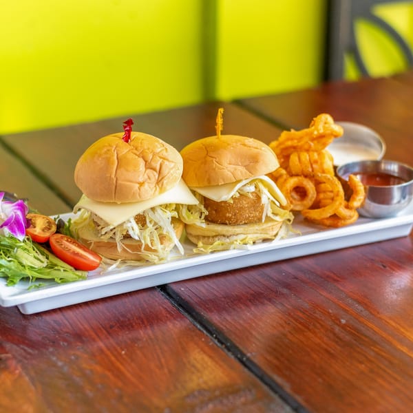 Two Indian-style sliders with cheese and shredded lettuce, curly fries, and dipping sauces on a white plate.